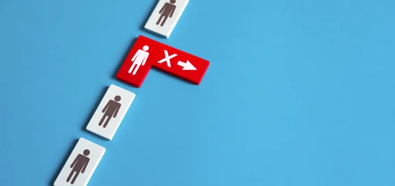 Ein Symbolbild Restrukturierung: Auf einem blauen Hintergrund liegen mehrere weiße Dominosteine mit schwarzen Personensymbolen. Einer der Steine ist rot, zeigt eine weiße Figur mit einem Pfeil nach rechts und einem 'X' und ist aus der Reihe entfernt.