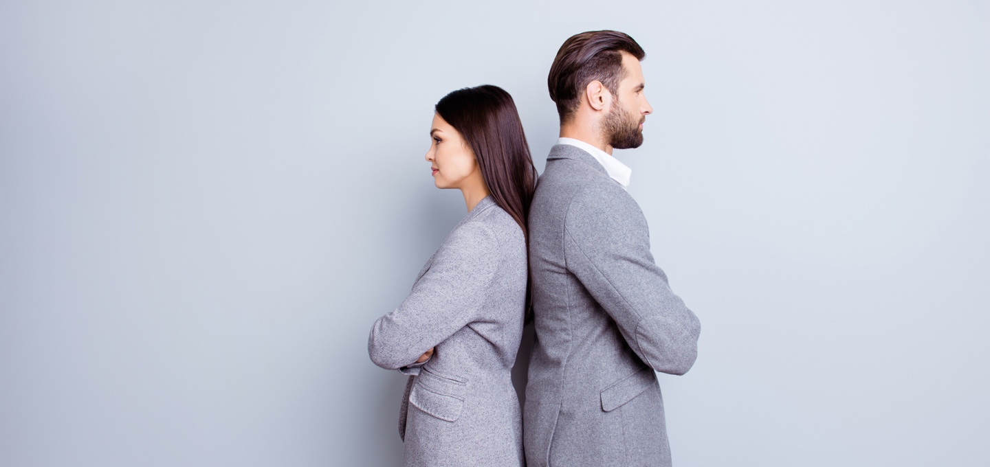 A woman and a man stand back to back with their arms crossed in front of a neutral background. Both are wearing grey business attire and looking in opposite directions - symbolising the gender pay gap.