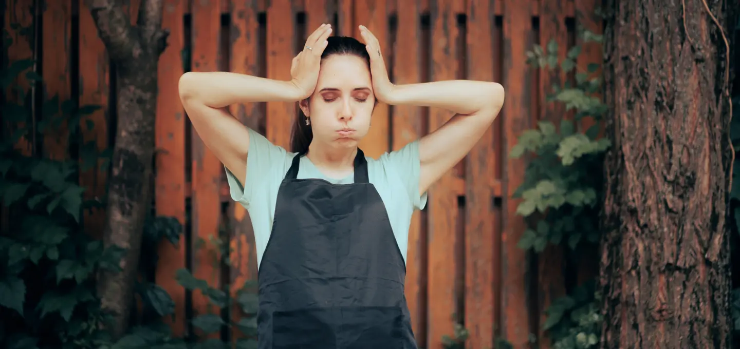 Eine Frau im Arbeitsoutfit steht mit Händen am Kopf und aufgeblasenen Wangen gestresst vor einem Holzzaun.