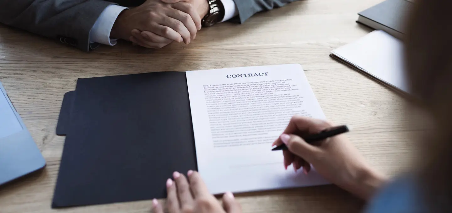 Zwei Personen sitzen an einem Tisch, eine davon unterschreibt einen Vertrag, der aufgeschlagen in einer schwarzen Mappe liegt.