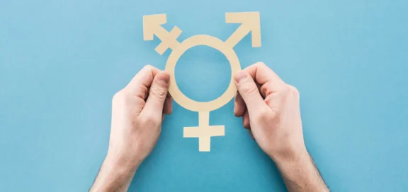 Zwei Hände halten ein ausgeschnittenes Transgender-Symbol auf blauem Hintergrund.
