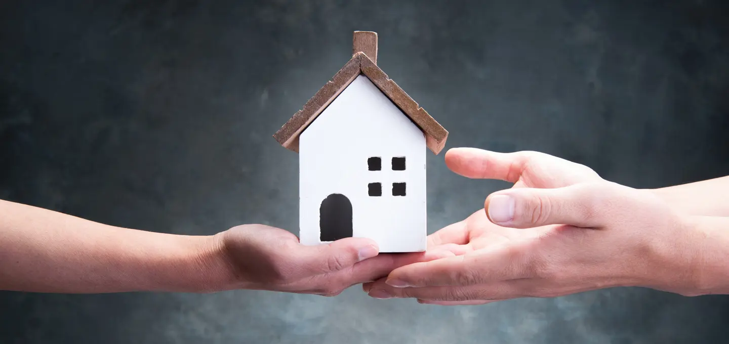 Zwei Hände übergeben symbolisch ein kleines weißes Modellhaus mit braunem Dach – Sinnbild für Immobilienübertragung und Nachfolgeplanung im Rahmen der Erbschaftsteuer 2025/26.