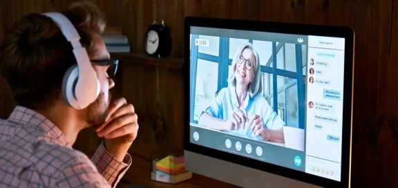 Ein Mann mit Kopfhörern sitzt vor einem Computerbildschirm und nimmt an einem Online-Videoanruf teil. Auf dem Monitor ist eine ältere Frau mit Brille zu sehen, die lächelt und spricht. Rechts im Bild ist ein Chatfenster mit Nachrichten der Teilnehmer sichtbar - Symbolisch für die Nichtigkeit von Online-Coaching-Verträgen