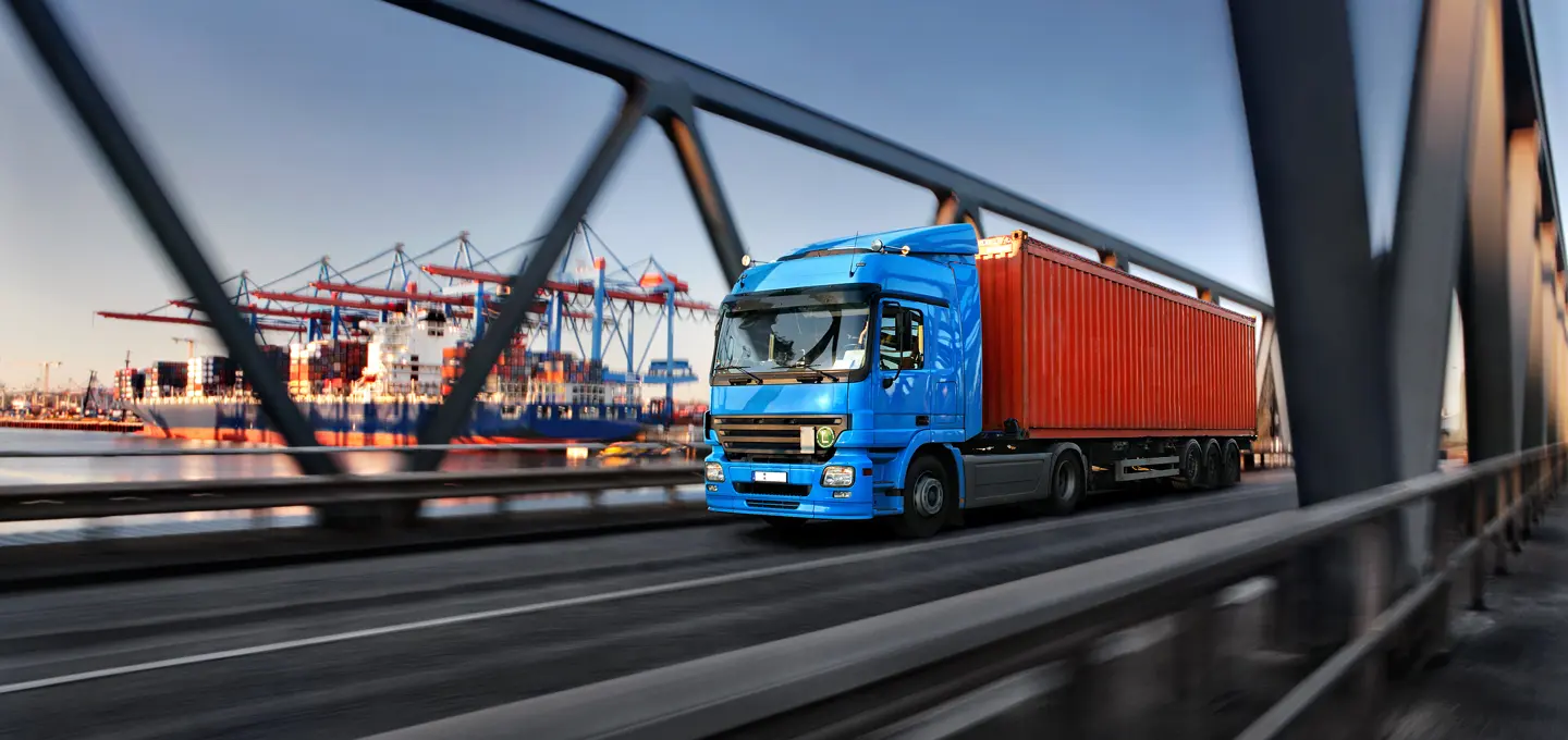 Blue lorry with red container drives over a bridge in a harbour, with container cranes and a cargo ship visible in the background.