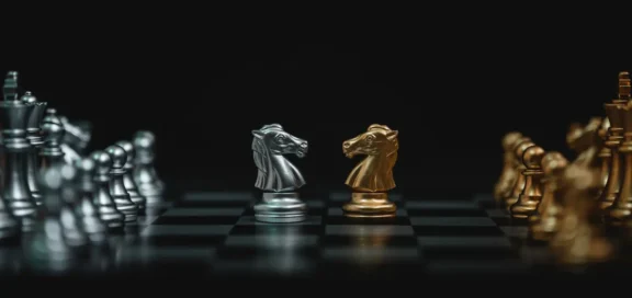 Nahaufnahme eines Schachbretts mit zwei gegenüberstehenden Springern in Silber und Gold im Zentrum, flankiert von silbernen und goldenen Schachfiguren im Hintergrund, vor einem dunklen, unscharfen Hintergrund – Symbloisch für die Prozessfinanzierung