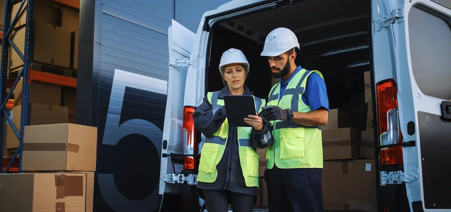 Symboldbild für Claim Management in der Logistik – Zwei Logistikmitarbeiter mit Schutzhelm und Warnweste prüfen auf einem Tablet Lieferdaten vor einem offenen Transporter mit Paketen im Lager.