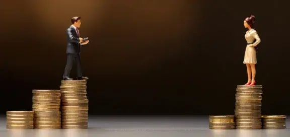 Zwei Miniaturfiguren stehen auf ungleich hohen Münzstapeln und blicken sich an. Der männliche Geschäftsmann steht links auf dem höchsten Stapel, während die Frau rechts auf einem niedrigeren Stapel steht, was den geschlechtsspezifischen Gehaltsunterschied (Equal Pay Verstoß) symbolisiert.