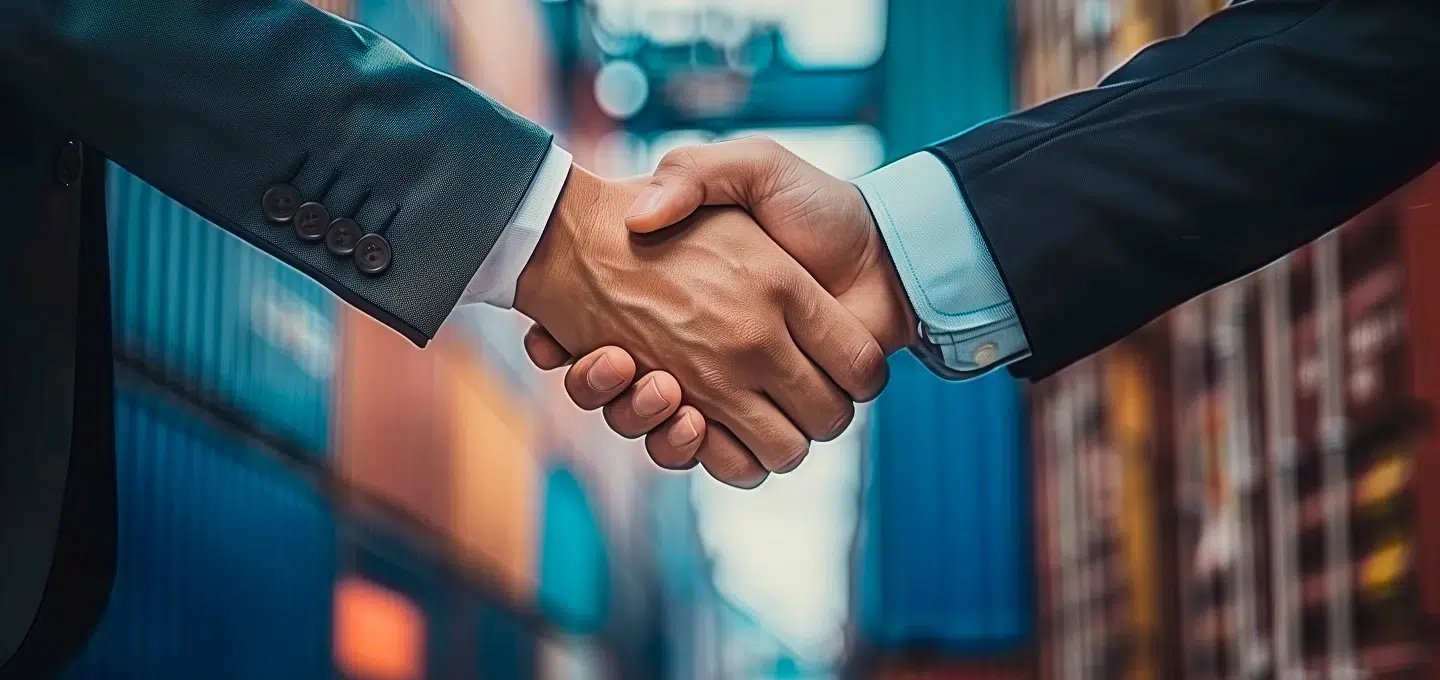 Nahaufnahme eines Händedrucks zwischen zwei Personen in Business-Anzügen vor einem unscharfen Hintergrund aus bunten Schiffscontainern – Symbolbild für internationale Lieferverträge