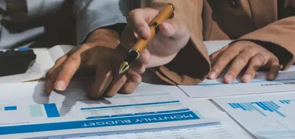 Unwirksame Nachrangklauseln – Nahaufnahme von Händen auf einem Schreibtisch, die Finanzdokumente prüfen. Im Vordergrund liegt eine Tabelle mit der Überschrift „Monthly Budget“ und blauen Diagrammen. Eine Person zeigt mit einem edlen Füllhalter auf die Daten.