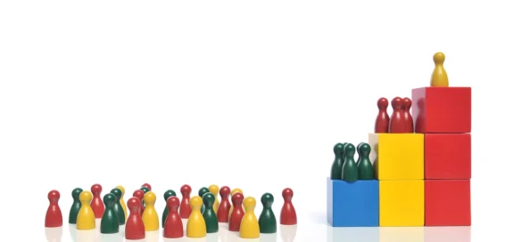 Spielfiguren in Rot, Gelb und Grün: Eine ungeordnete Gruppe steht links, während sich rechts drei Gruppen hierarchisch auf bunten Stufen aus Holzwürfeln anordnen – Sinnbild für die Betriebsratsgründung ohne inländischen Hauptbetrieb