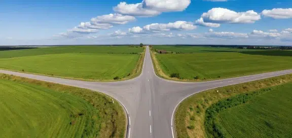 Kreuzung einer Landstraße inmitten grüner Felder unter blauem Himmel als Symbol für die Wahl des richtigen Sanierungswegs in der Unternehmenskrise